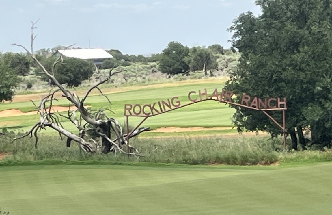 An arched metal sign reading ROCKING CHAIR RANCH stands at the edge of a green field, next to a leafless, twisted tree&mdash;hinting at classic cattle country&mdash;with trees and a white-roofed building in the background.