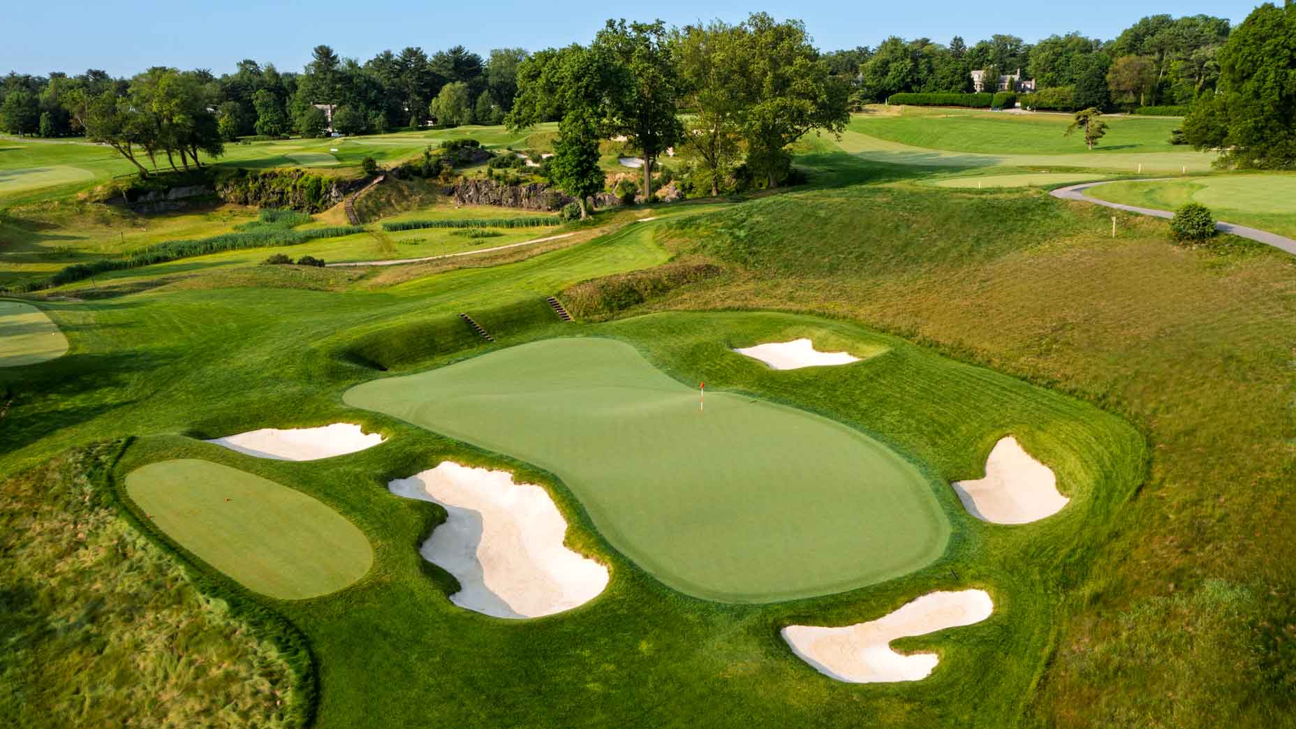 A lush golf course at Merion featuring a green surrounded by several sand bunkers, with manicured fairways, trees, and a winding path in the background&mdash;an inviting scene where many have flown too close to the sun.