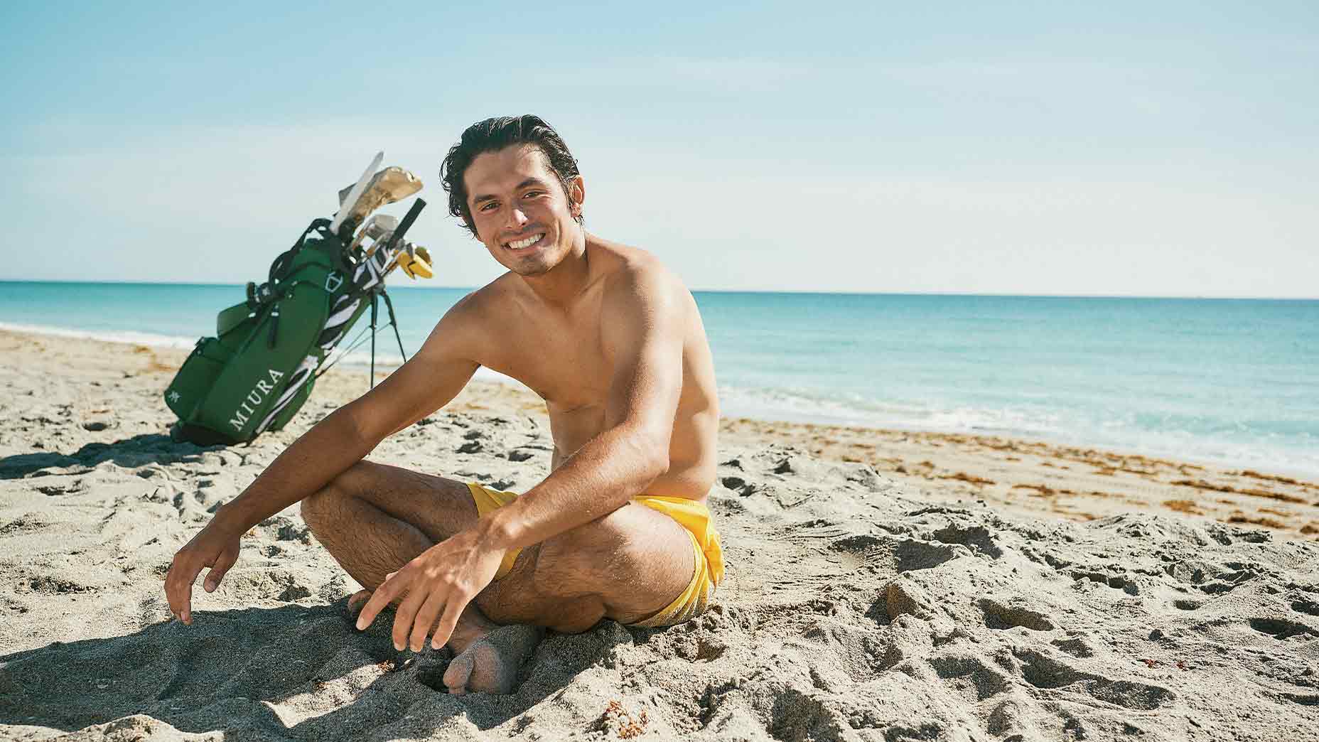 A man in yellow swim trunks sits smiling on a sandy beach near the ocean, with a green golf bag and clubs behind him under a clear blue sky&mdash;a scene reminiscent of Fat Perez, one of golf&rsquo;s digital boom online creators, merging relaxation and the love of the game.