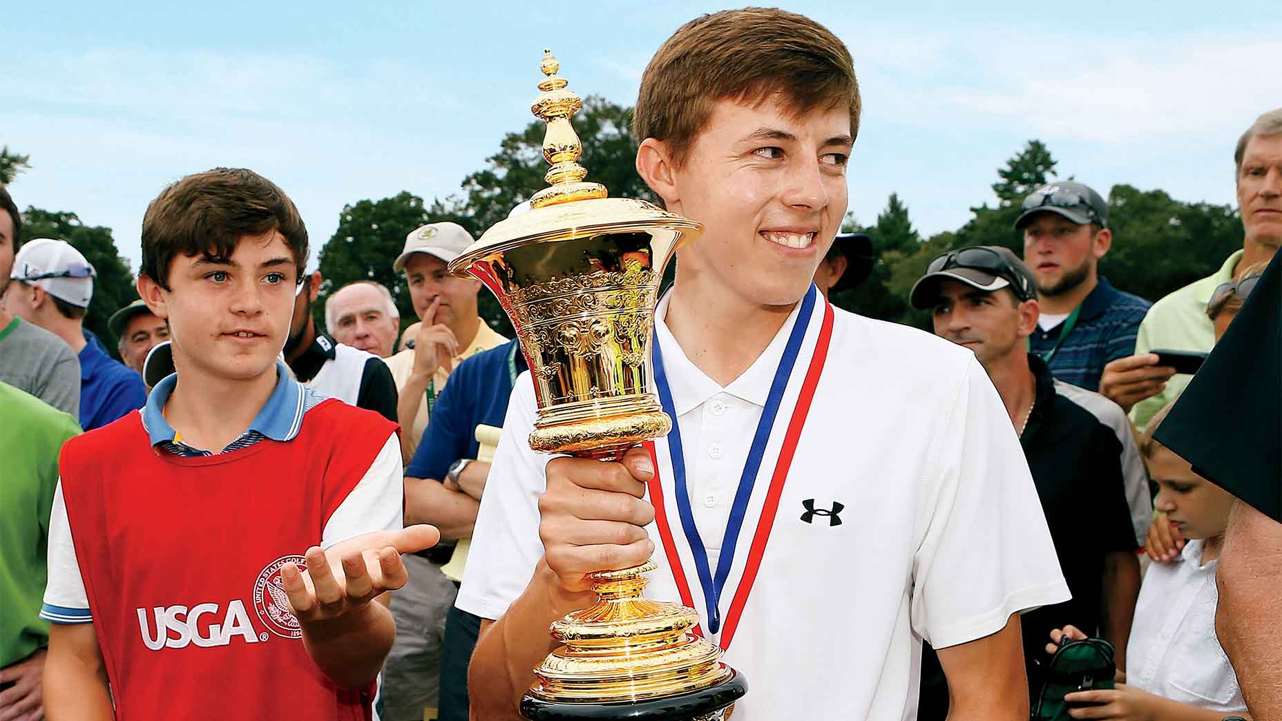 A young golfer in a white shirt holds a large gold trophy and wears a medal, smiling, surrounded by people outdoors, reminiscent of the celebrations seen at the FedEx Cup playoffs. Another young person in a red vest stands nearby, perhaps offering congratulations just as Alex Fitzpatrick might among fellow competitors.
