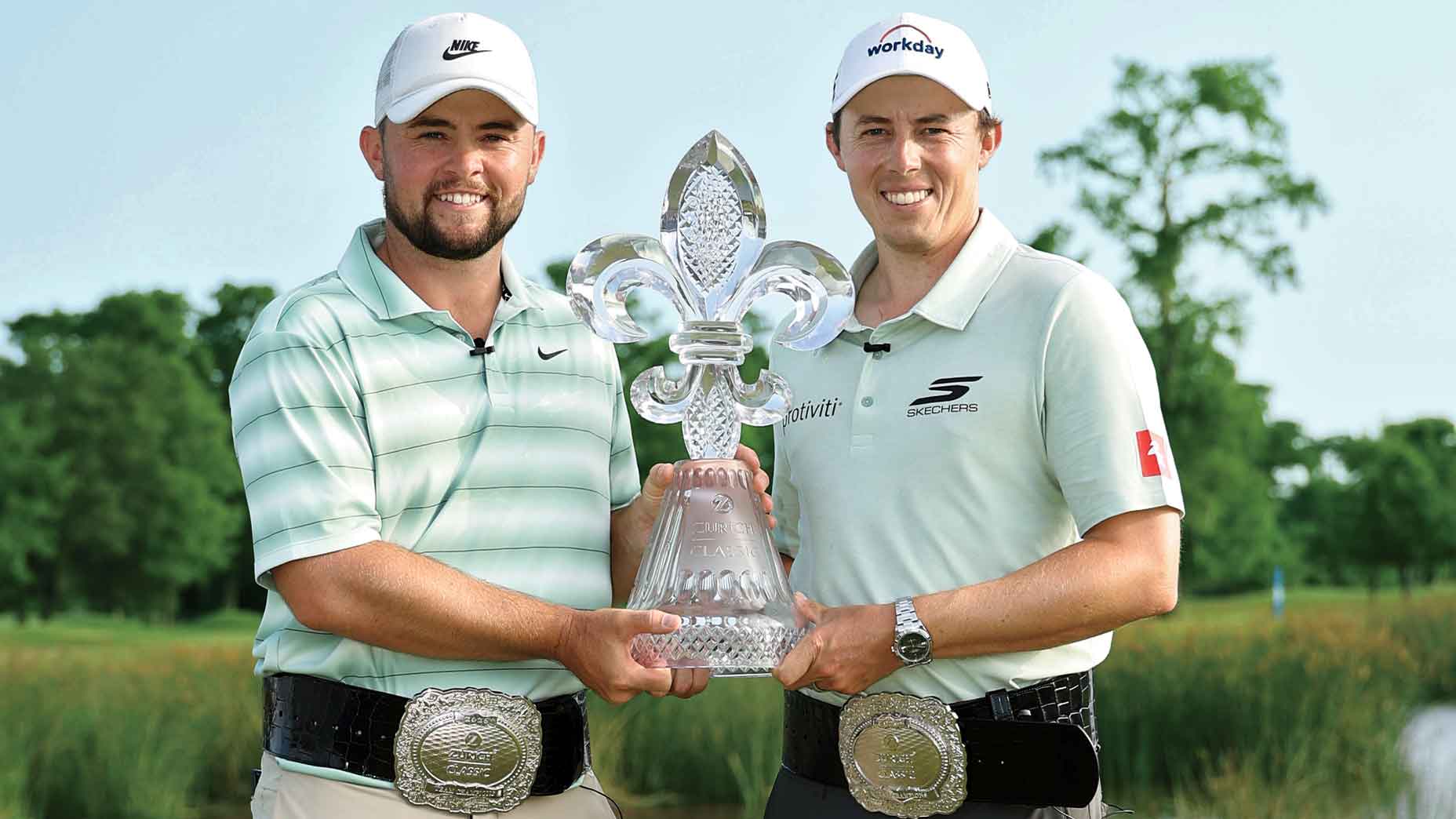 Two golfers, both in light shirts and wearing caps, stand outdoors holding a large trophy shaped like a fleur-de-lis. They are smiling and wearing large decorative belt buckles, reminiscent of those seen at major tournaments such as the FedEx Cup playoffs. Trees and grass are visible in the background.