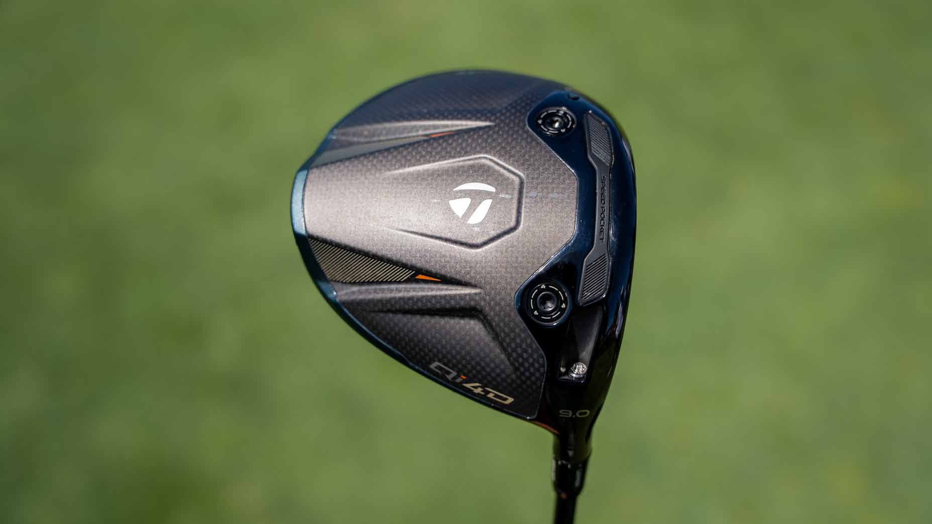 Close-up of a TaylorMade golf driver clubhead with a black and silver design, featuring logos and adjustment screws, set against a blurred green background&mdash;a setup reminiscent of equipment found in Tour Report features or used by professionals like Tommy Fleetwood.