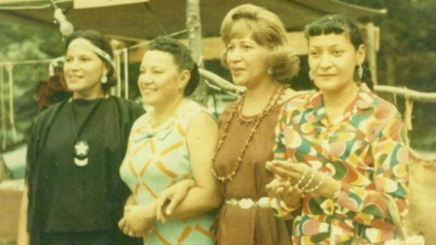 Four women stand closely together outdoors in Shinnecock Hills, smiling and holding hands. Dressed in colorful, patterned clothing and jewelry, they share a family bond. Trees and a wooden structure appear in the vintage photo&rsquo;s background.