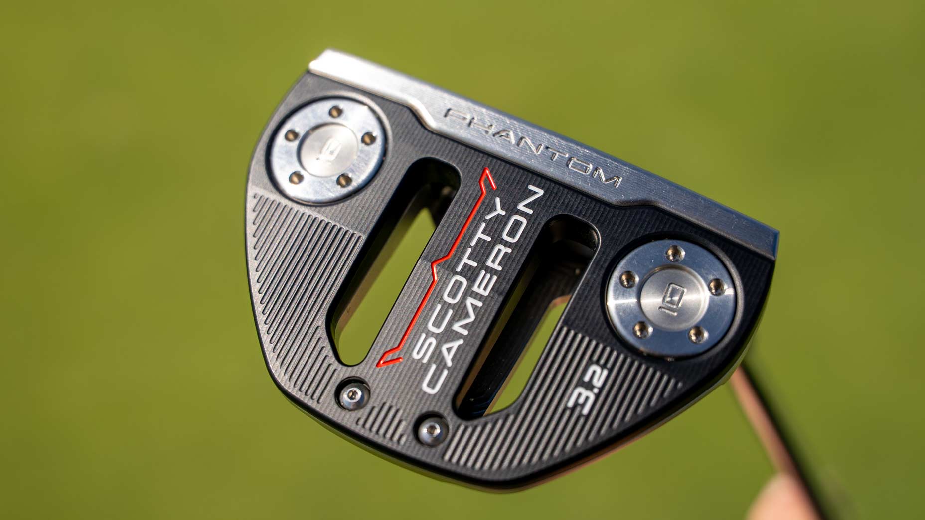 Close-up of a Scotty Cameron Phantom mallet 2.5 putter head, featuring new shapes in black and silver details, red and white branding, and visible screws, set against a blurred green background.