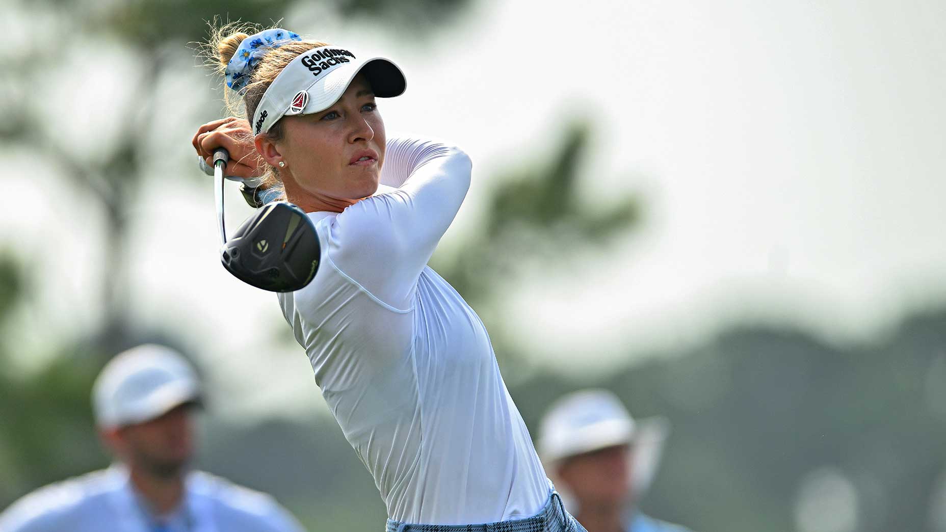 Nelly Korda in a white long-sleeve shirt and visor follows through on her swing at the U.S. Women's Open, holding a driver, with blurred spectators and trees in the background.