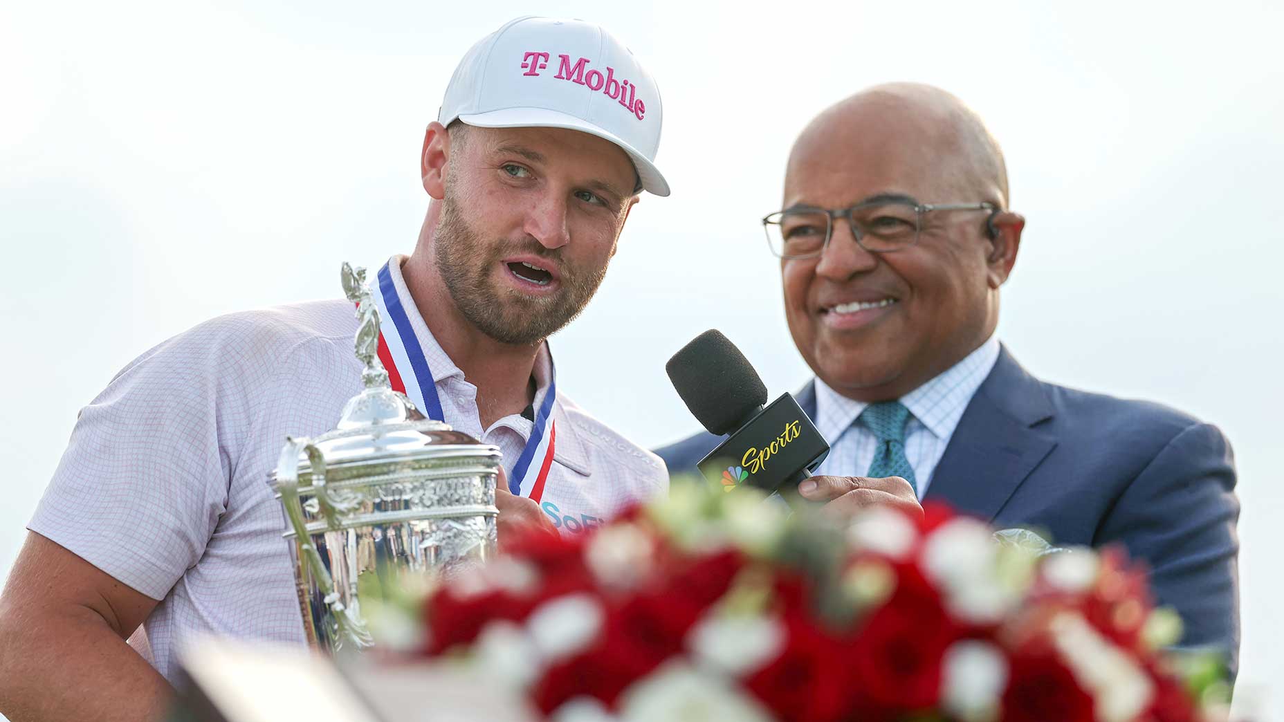 A man in a T-Mobile cap, identified as U.S. Open winner Wyndham Clark, is being interviewed by a smiling broadcaster in a suit holding an NBC Sports microphone, with a bouquet of red and white flowers in the foreground.