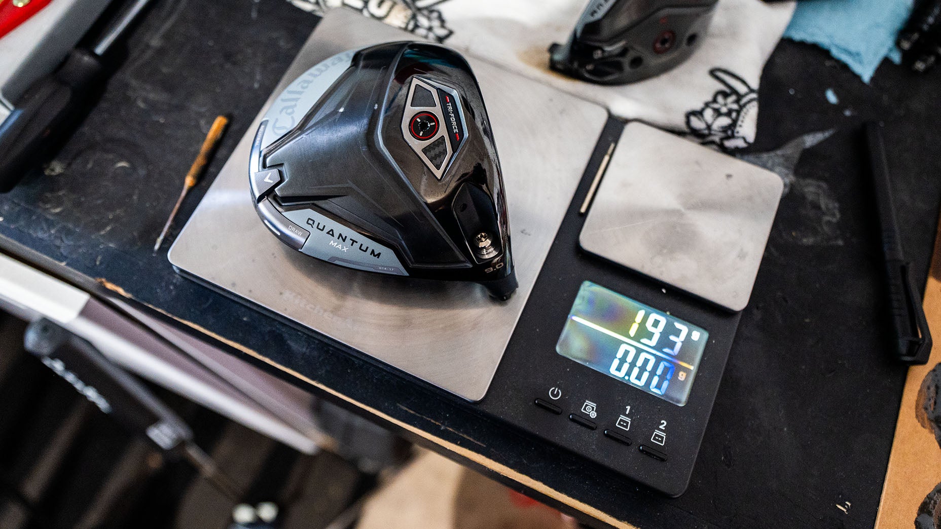 A black golf club head labeled Quantum rests on a digital scale showing 193.7 grams, while nearby golf shafts and tools hint at the precision of a dedicated manufacturer&rsquo;s workspace.