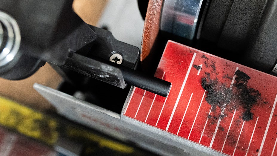 Close-up of a cutting tool slicing through a black golf shaft tip on a red, gridded cutting surface, with dark debris scattered on the board.