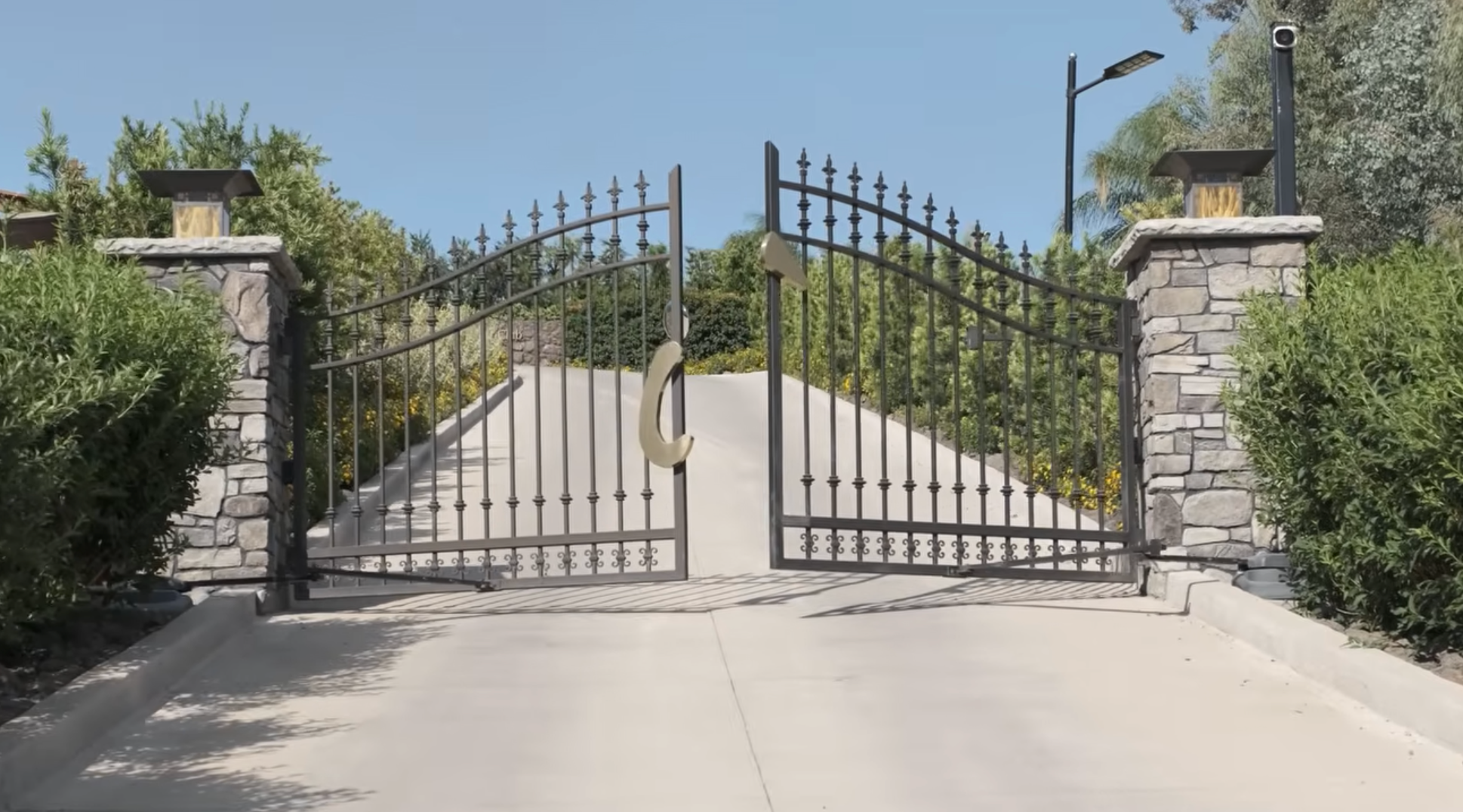 A closed wrought iron gate with stone pillars, marked by a sign for The Institute, blocks a concrete driveway leading uphill, surrounded by green shrubs and trees under a clear blue sky.