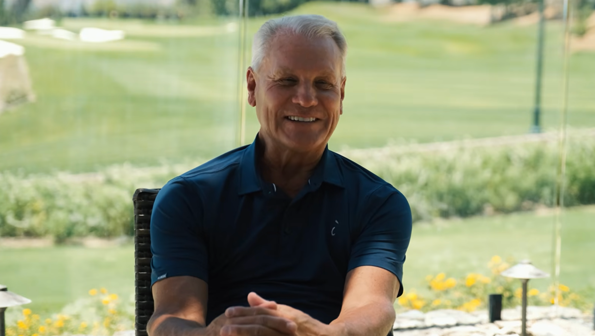 A smiling older man with short gray hair sits outdoors on a chair, hands clasped, wearing a dark polo shirt. Behind him, the lush golf course and greenery of The Institute are visible in the sunny background.