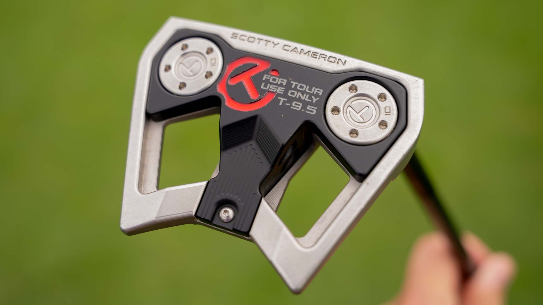Close-up of a new putter: the Scotty Cameron T9.5 golf putter head with For Tour Use Only text, silver and black color, and screws visible, held outdoors with green grass in the background. Featured in a recent Tour Report and similar to the model used by Brooks Koepka.