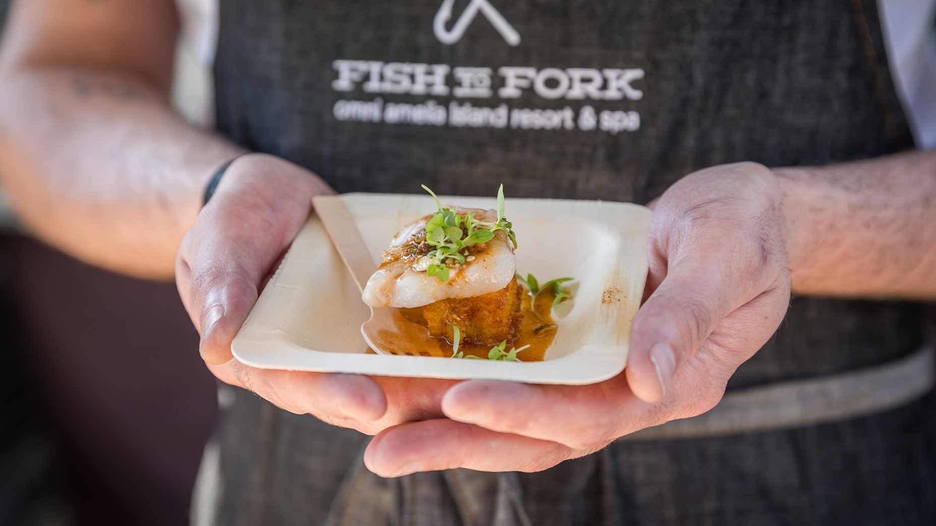 A person wearing a Fish to Fork apron holds a small wooden plate with gourmet seafood topped with microgreens, and a wooden fork resting on the plate.