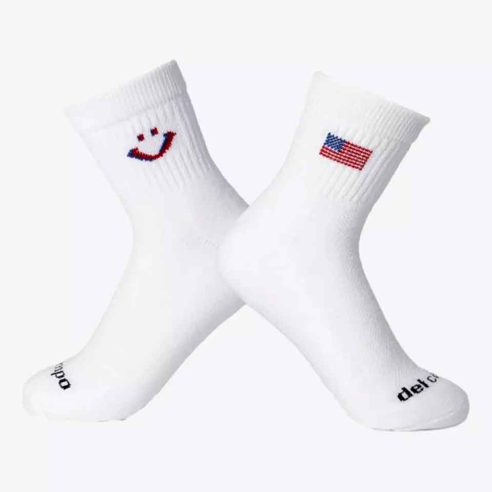 Two white socks crossed at the ankle&mdash;one with a small American flag design, the other with a smiley face made from red and blue stitches. Both feature black text near the toes, making them a fun Father's Day gift.