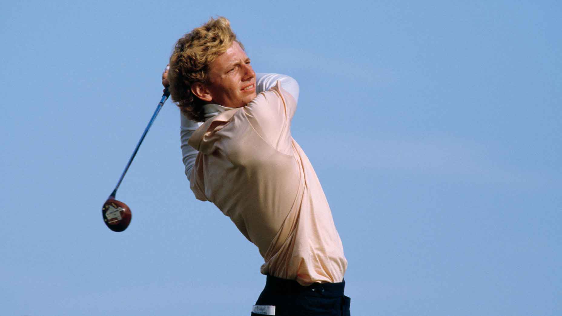 A person with curly blond hair swings a golf club outdoors under a clear blue sky, looking into the distance after taking a shot—a scene reminiscent of Brad Faxon during the 1985 Honda Classic.