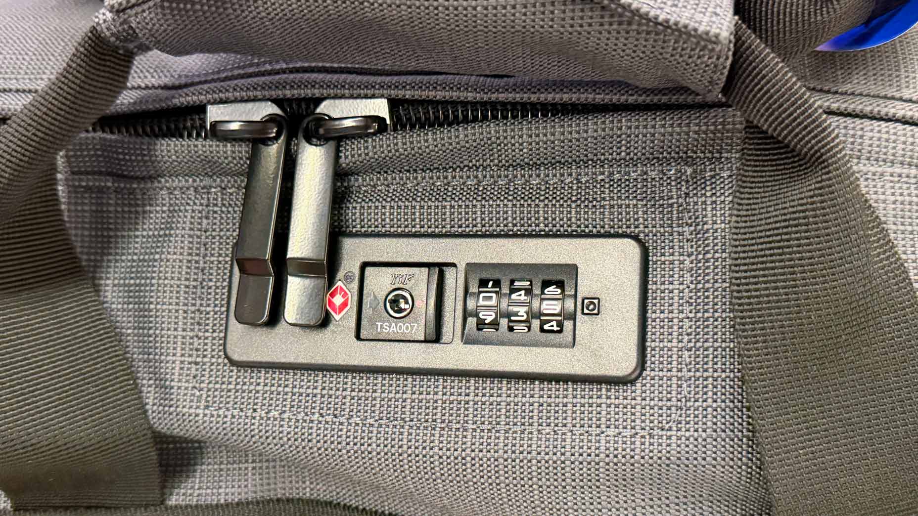 Close-up of a gray fabric travel bag with a built-in TSA-approved combination lock, showing zippers secured in the lock and a visible three-digit code dial.