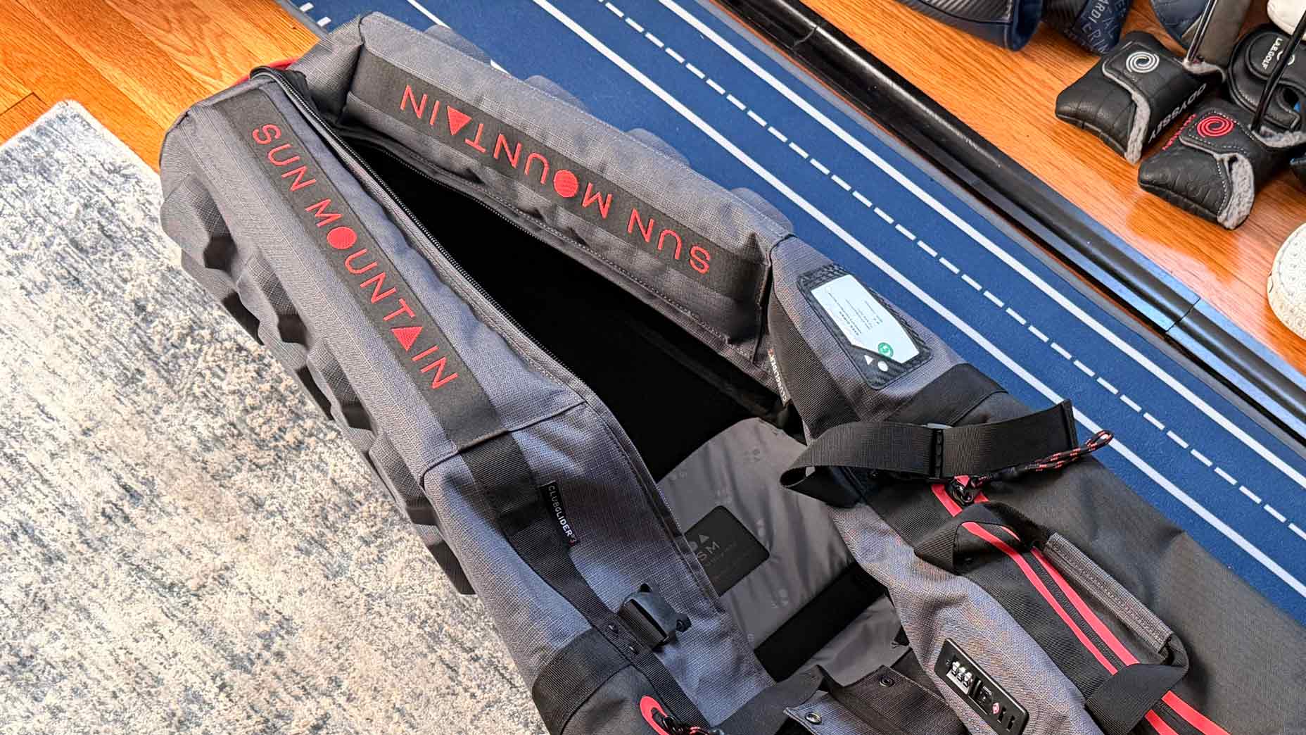 A gray Sun Mountain travel bag with red lettering lies open on a blue striped mat, showing its interior and zipper details. Golf gear is visible in the background.