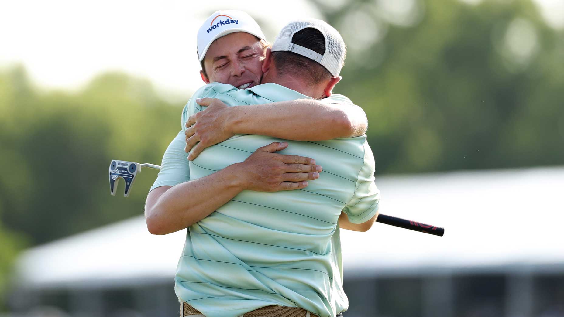 Alex and Matt Fitzpatrick embrace tightly on a golf course, one holding a golf club. The background is blurred with greenery and a structure visible&mdash;Matt Fitzpatrick celebrates on the PGA Tour.