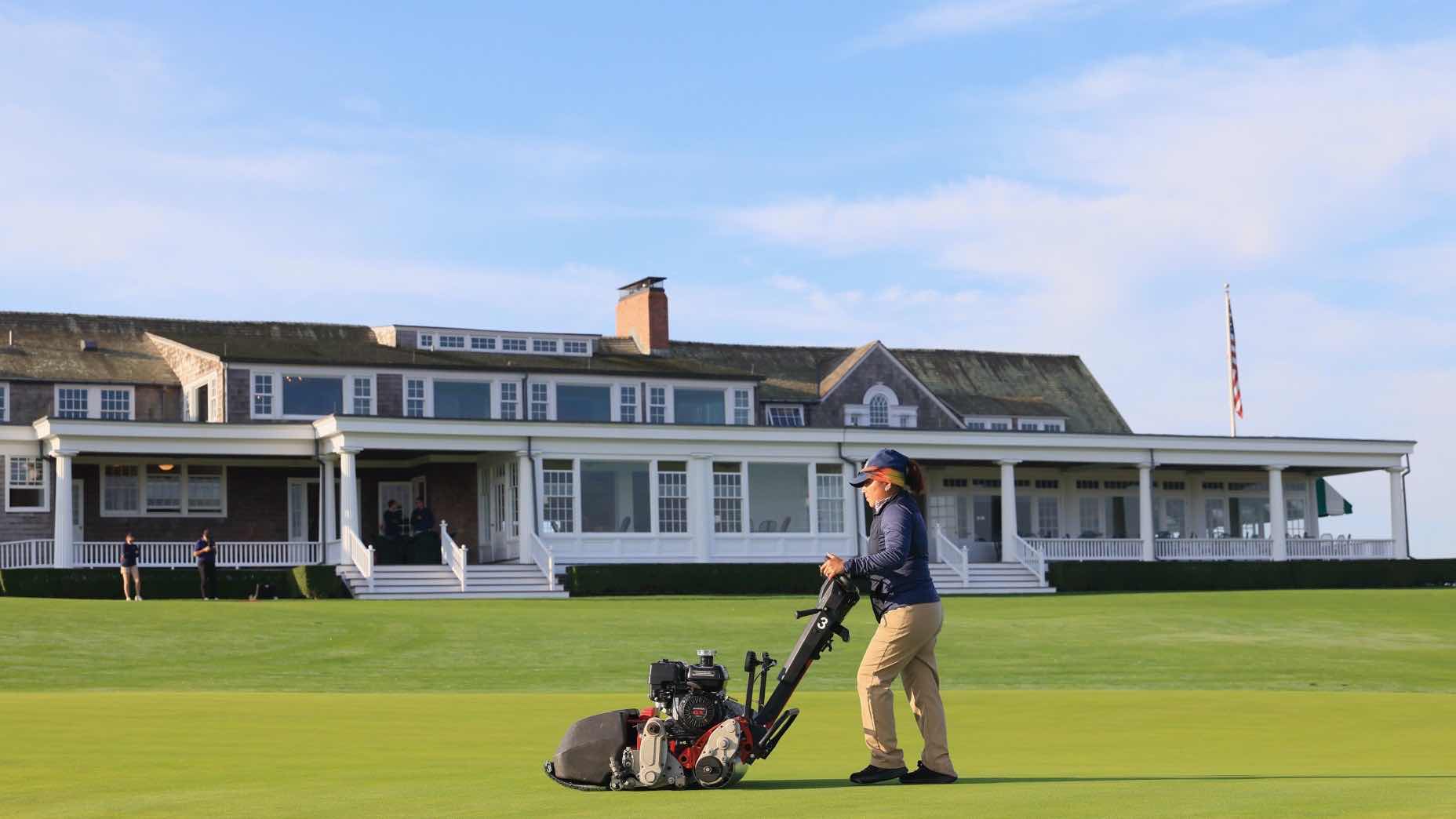 A person operates a lawn mower on a well-manicured golf course green, with a large clubhouse building and several people in the background under a blue sky.