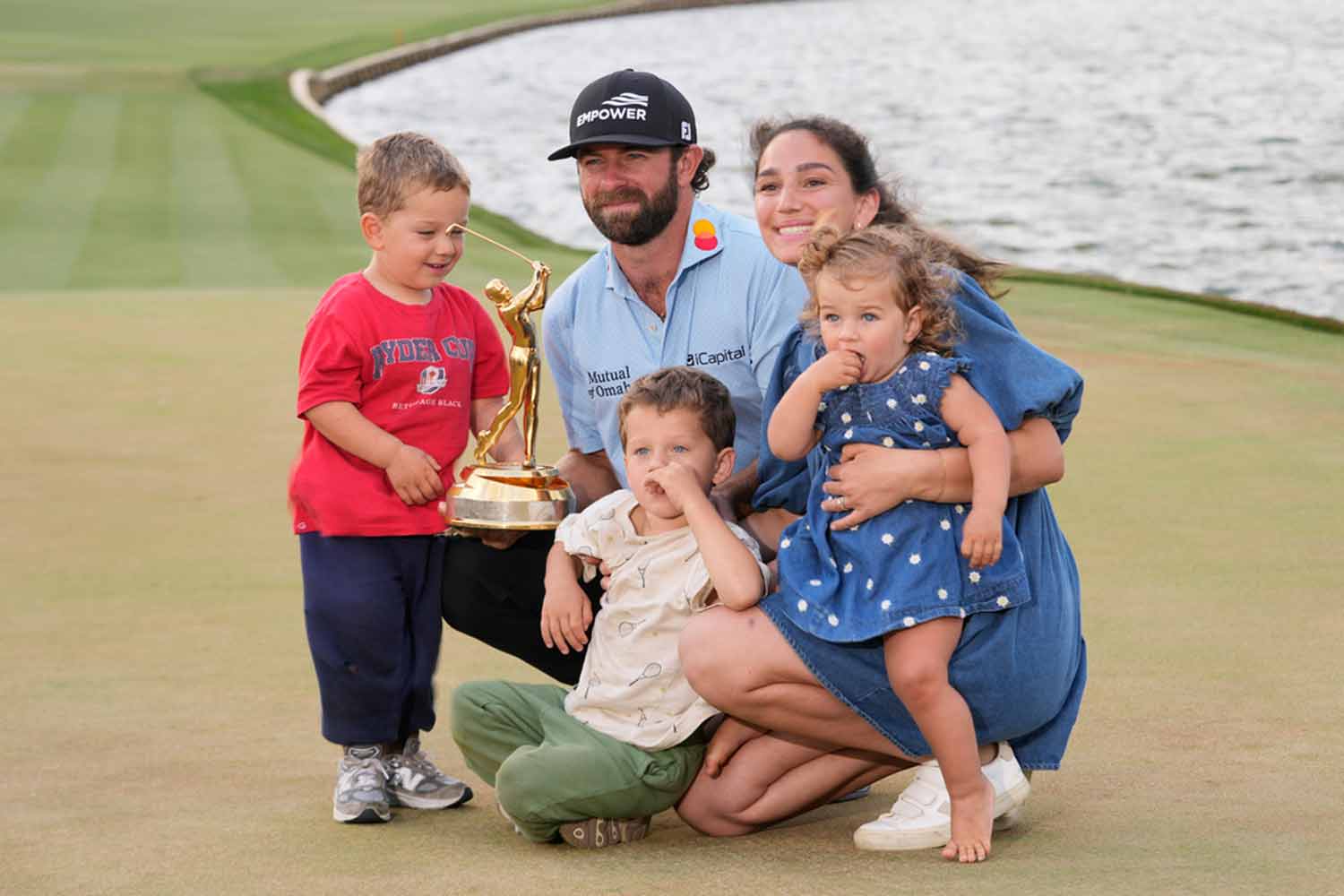 A golfer kneels on a green holding a trophy, surrounded by a woman and three young children. They all smile and pose together with a body of water visible in the background.
