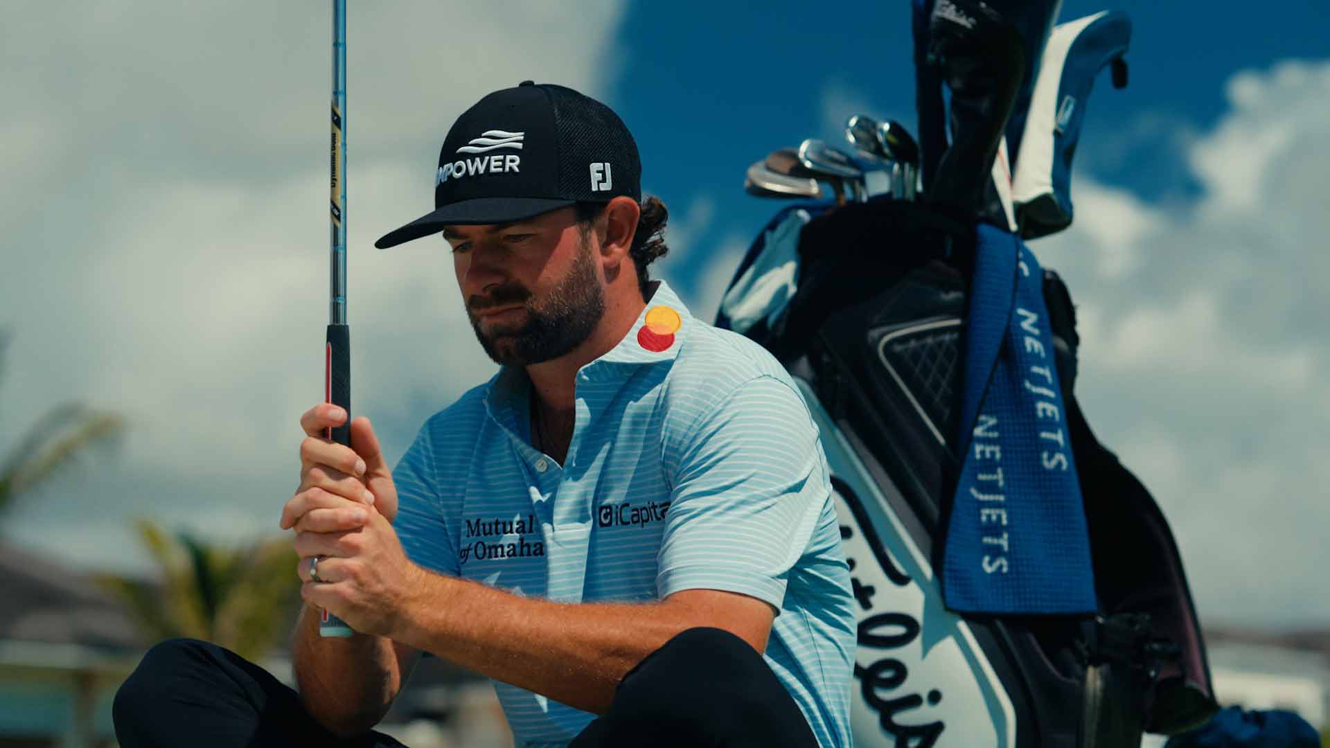 A male golfer wearing a cap and blue shirt squats on a golf course, holding a club. There&rsquo;s a golf bag and towel beside him, with palm trees and blue sky in the background.