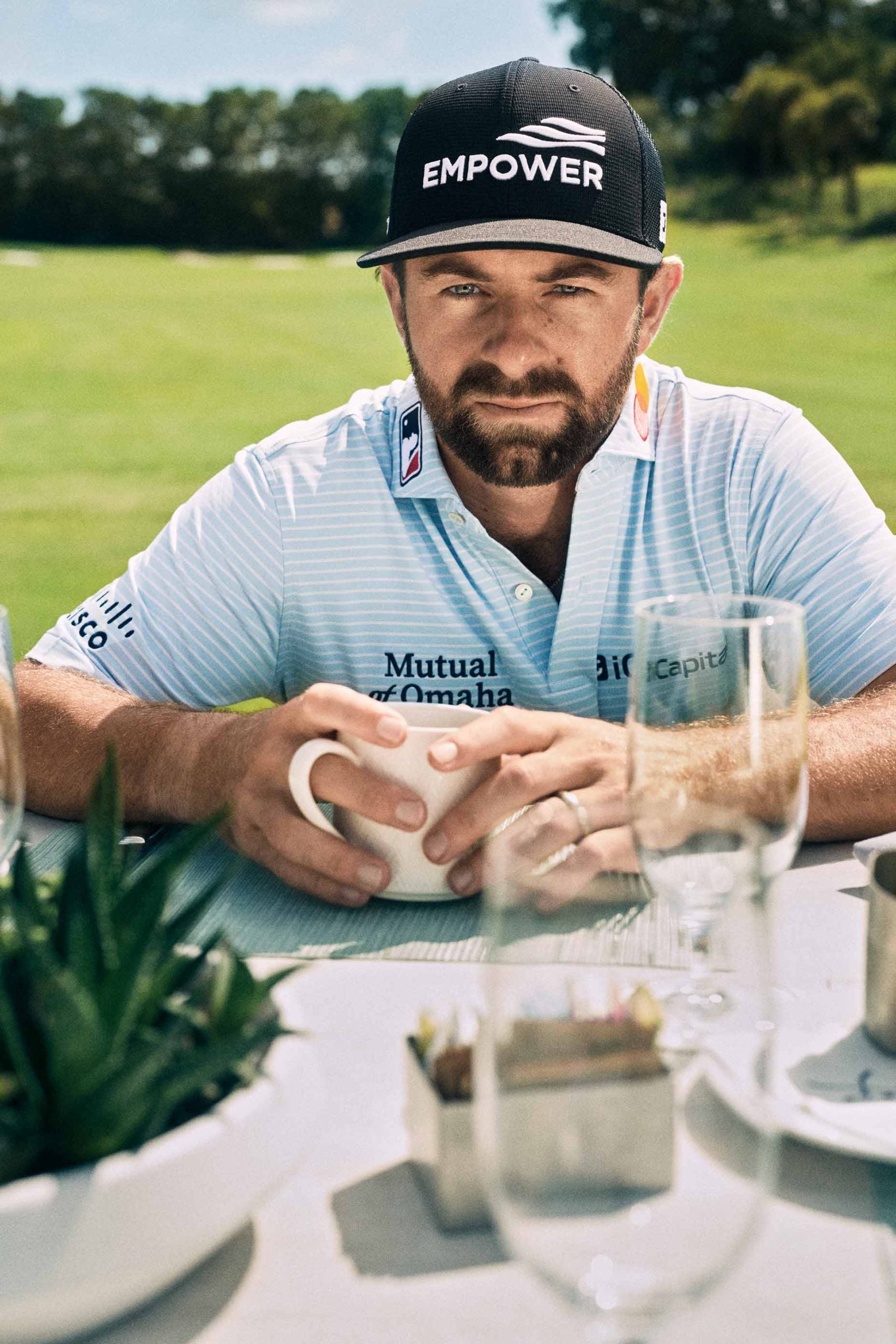 A man in a light blue striped golf shirt and black EMPOWER cap sits at an outdoor table holding a white mug, with golf course greenery in the background and glassware on the table in front of him.