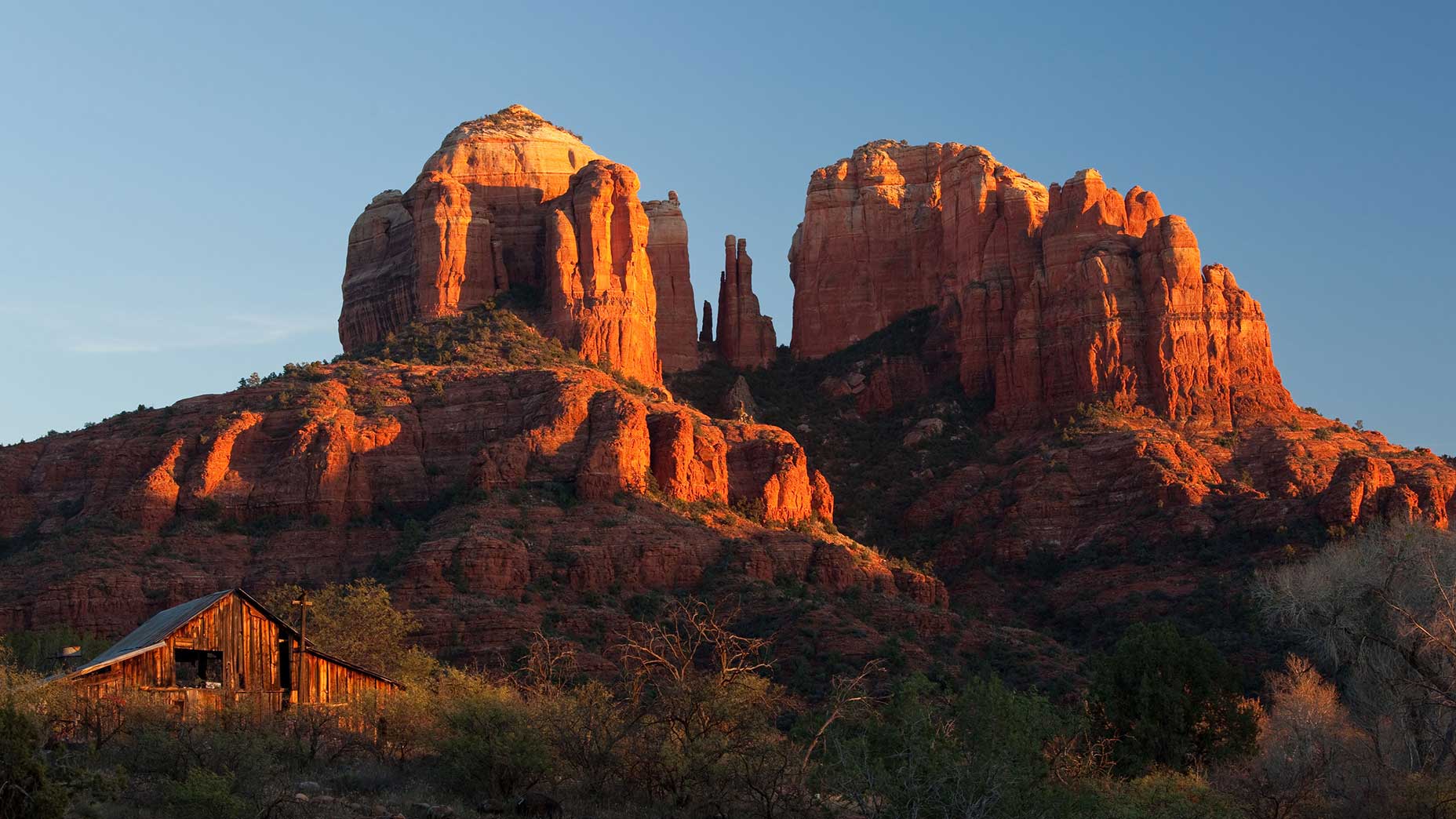 Red rock formations glow in the evening sun behind an old wooden barn, surrounded by green shrubs and bushes under a clear blue sky.