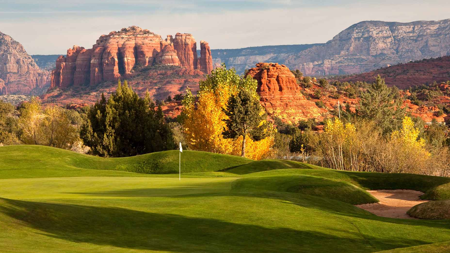 A lush green golf course sits in the foreground with a flag on the green, surrounded by trees and dramatic red rock formations under a blue sky in the background.
