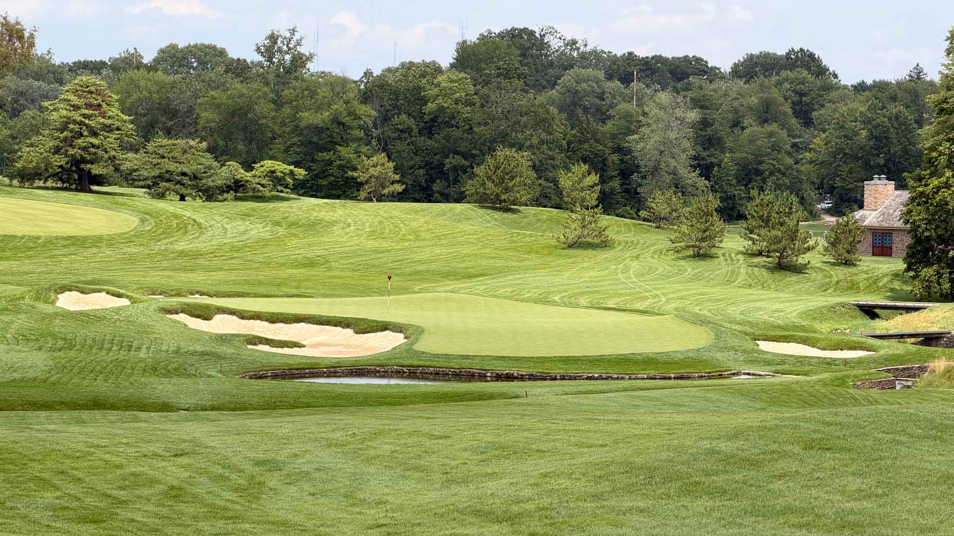 A well-maintained golf course with sand bunkers, a small pond, and a green surrounded by trees and a building in the background under a partly cloudy sky evokes the classic setting of Merion, home to historic moments like the U.S. Amateur. A lone golf bag stands near the fairway, ready for another memorable round on this storied layout.