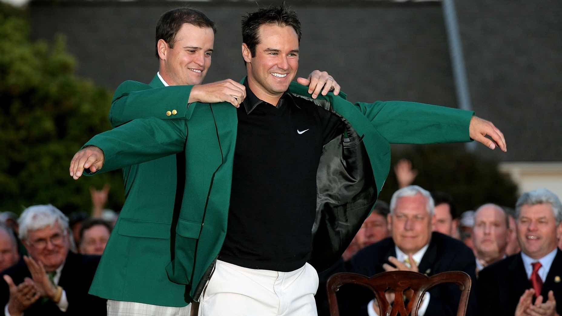 A smiling man is helped into a green jacket by Trevor Immelman, in front of a seated crowd outdoors. Both men appear happy, and the scene suggests a celebratory or award-giving moment.