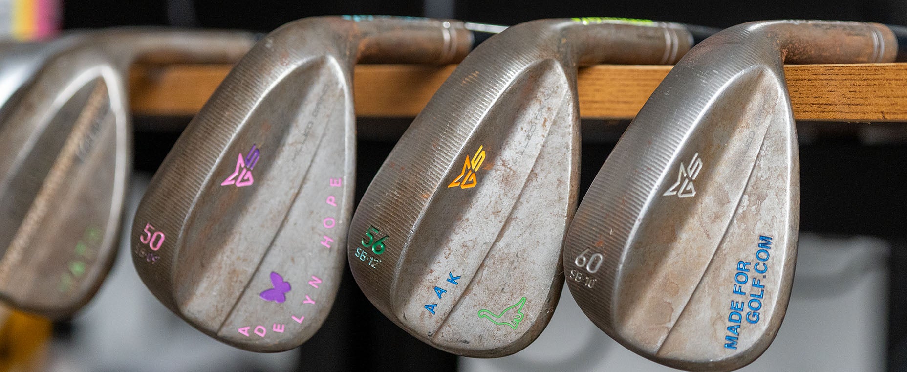 Three metal golf wedges with engraved, colorful text and initials rest side by side on a wooden rack. The clubs display numbers, symbols, and personalized engravings in pink, green, yellow, and blue&mdash;crafted to be your perfect wedges for a Fully Fit 2026 game.