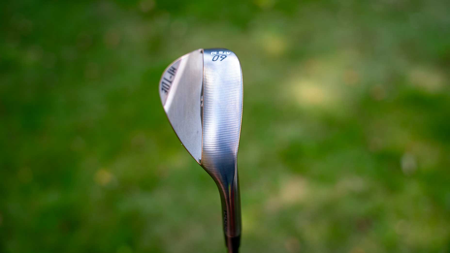 A close-up of a forged TaylorMade Hi-Toe wedge club, angled to show the shiny metal face and 60 loft marking, with a blurred green grass background.
