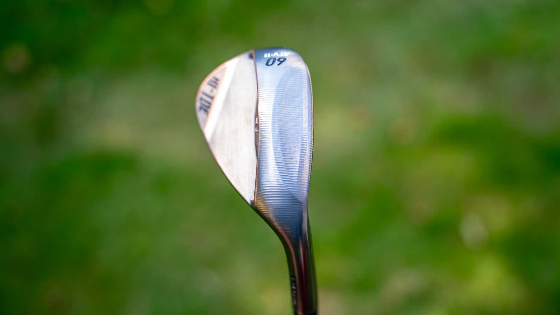 A close-up of a TaylorMade Hi-Toe wedge with a shiny forged metal finish, marked 60, standing upright against a blurred green grassy background.