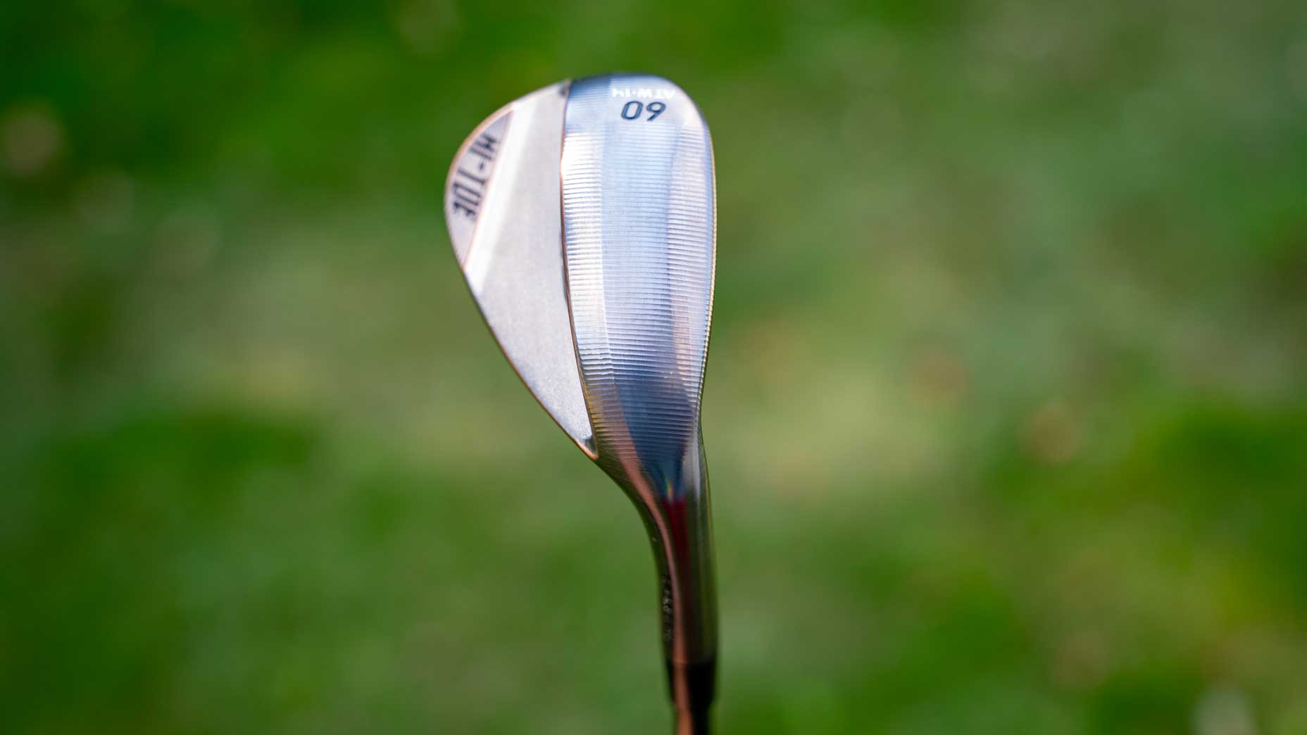A close-up of a forged Hi-Toe wedge from TaylorMade, showing the grooved clubface and the number 60 on its top, set against a blurred green background.