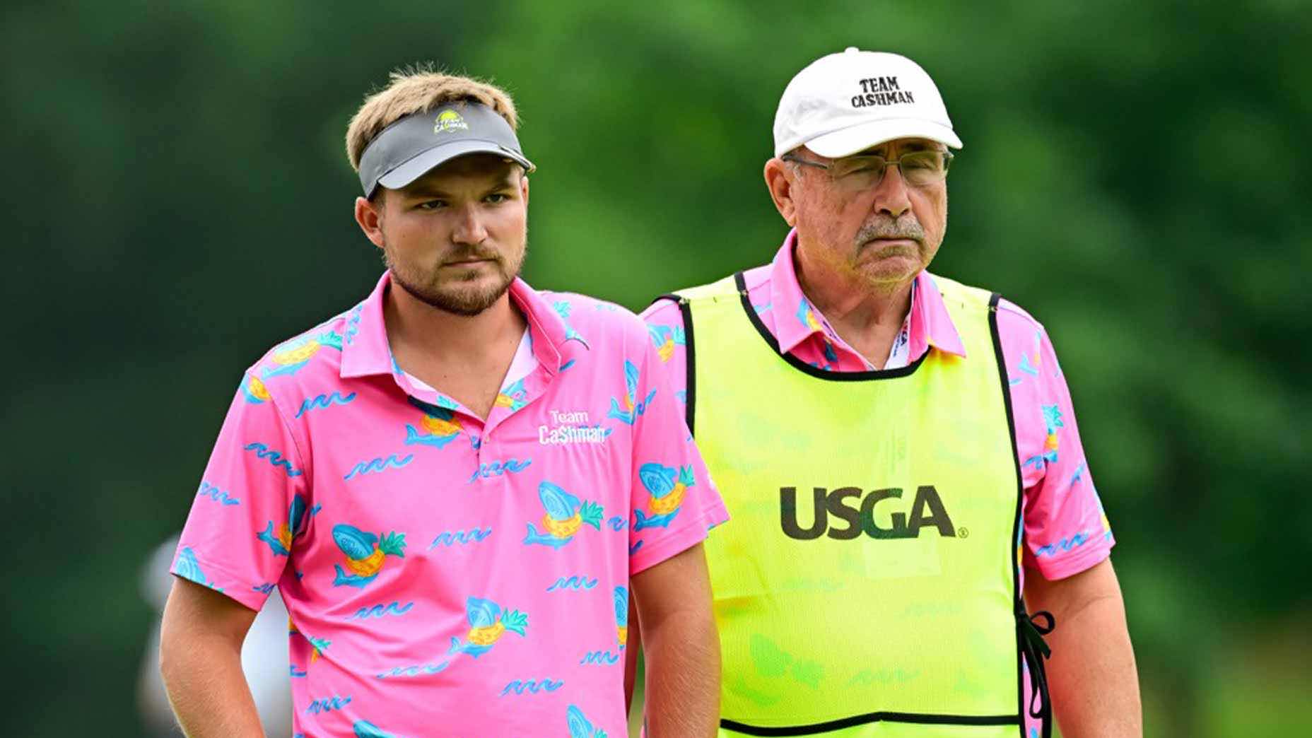 Two men stand side by side outdoors, both wearing matching bright pink shirts with colorful patterns. The man on the right, possibly a blind golfer's grandfather, also wears a white cap and a yellow vest labeled USGA. The background is blurred with greenery.