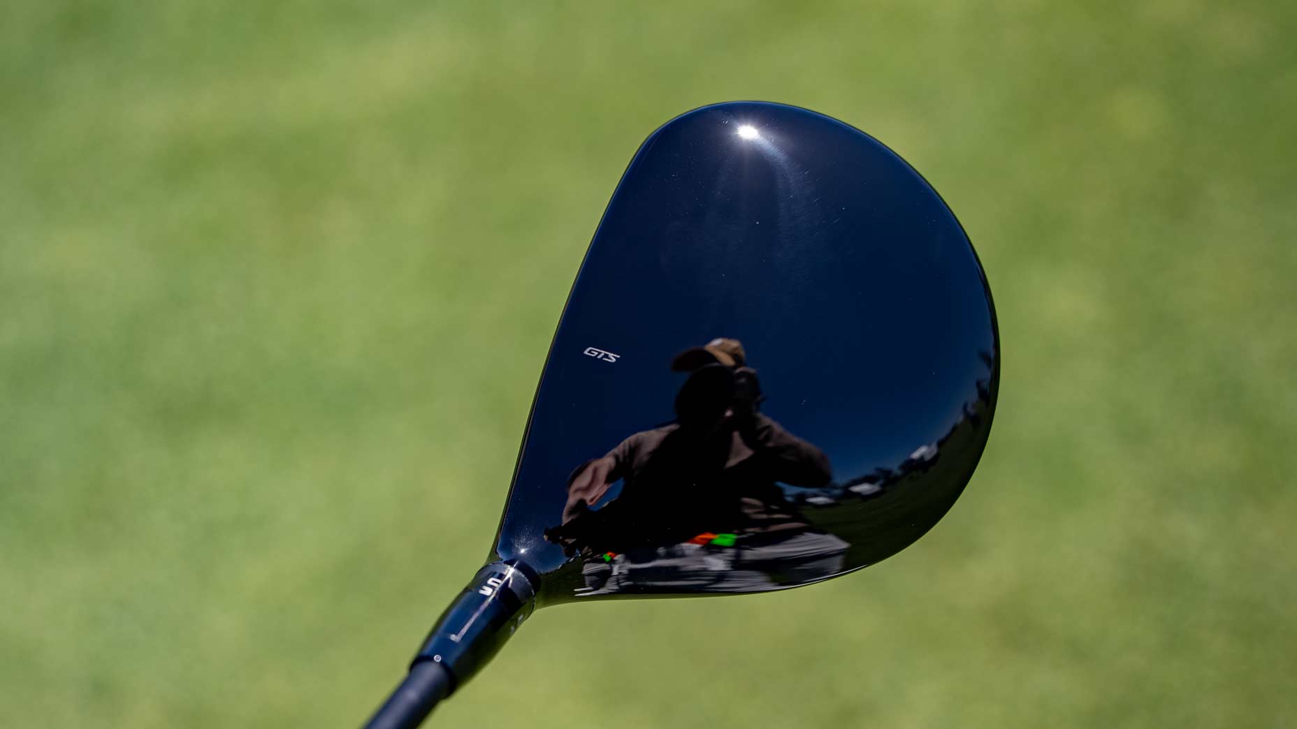 Close-up of a black Titleist GTS300 Mini Driver head reflecting a person with a hat holding a camera, outdoors on a green grassy field.
