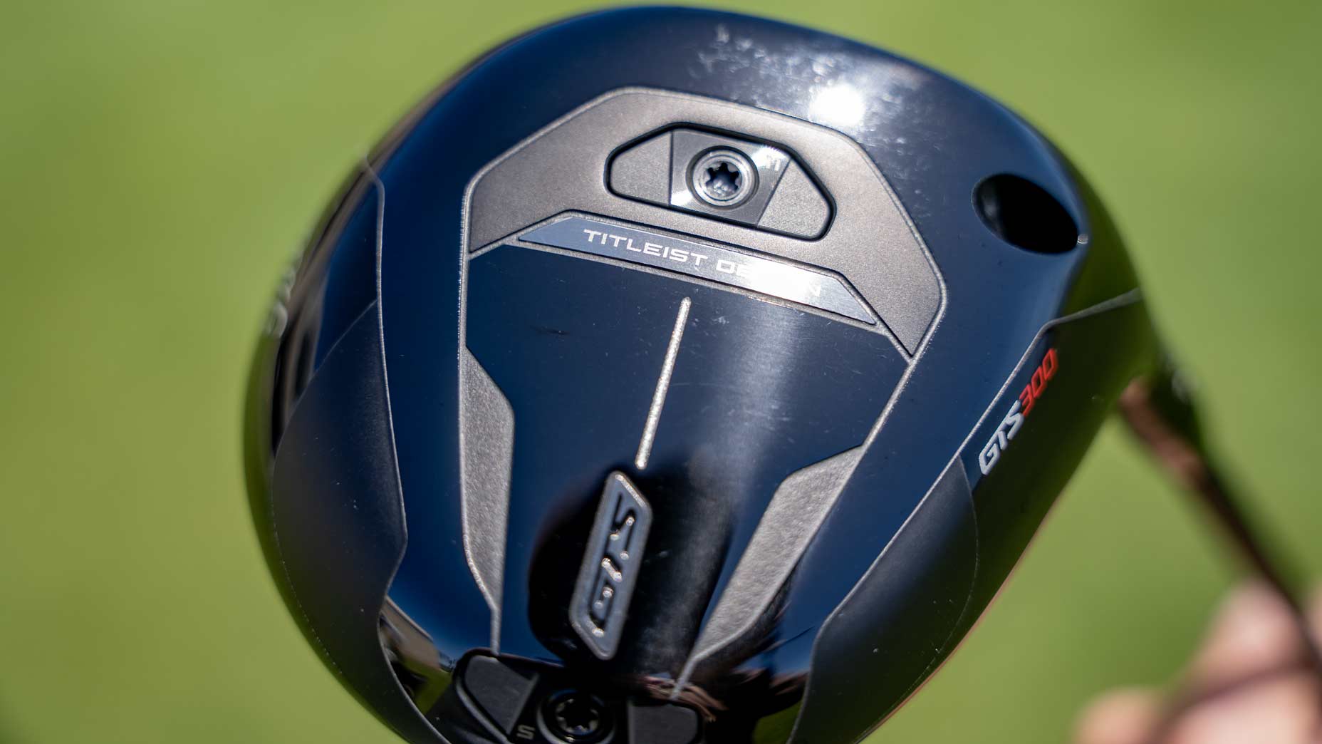Close-up of a black Titleist GTS300 golf club head with &ldquo;TS2&rdquo; and &ldquo;Titleist&rdquo; branding, screws, and sleek silver accents, highlighting the Mini Driver design, set against a blurred green background.