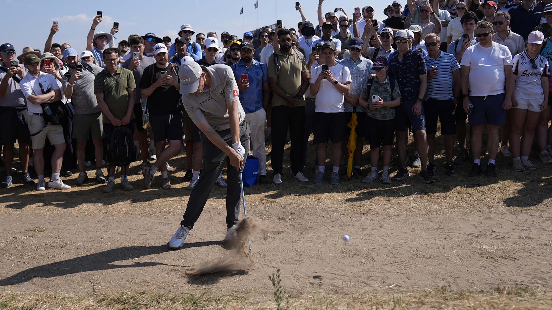 A golfer swings at a ball from a sandy patch as a large crowd watches closely, many filming with phones under a clear sky.