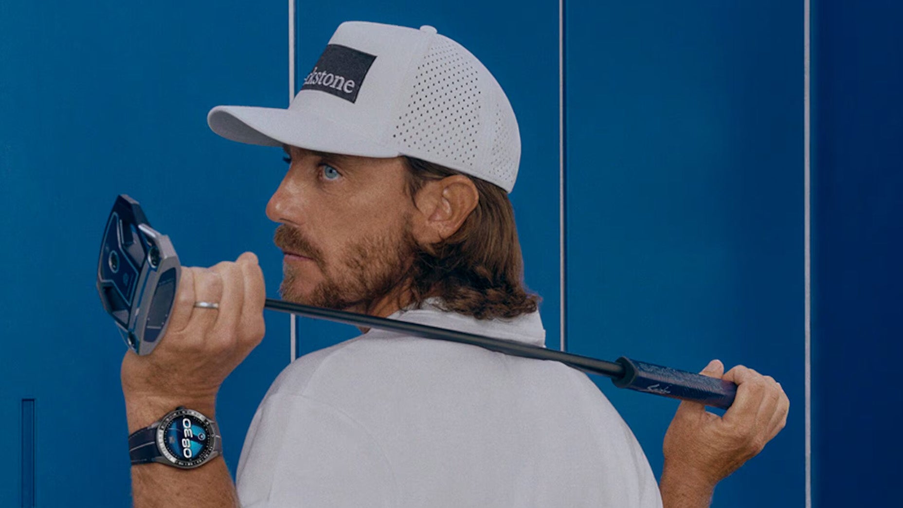 A man wearing a white cap and shirt holds a golf club over his shoulder. He is looking back over his left shoulder. He also wears a smartwatch on his left wrist. Blue lockers are visible in the background.