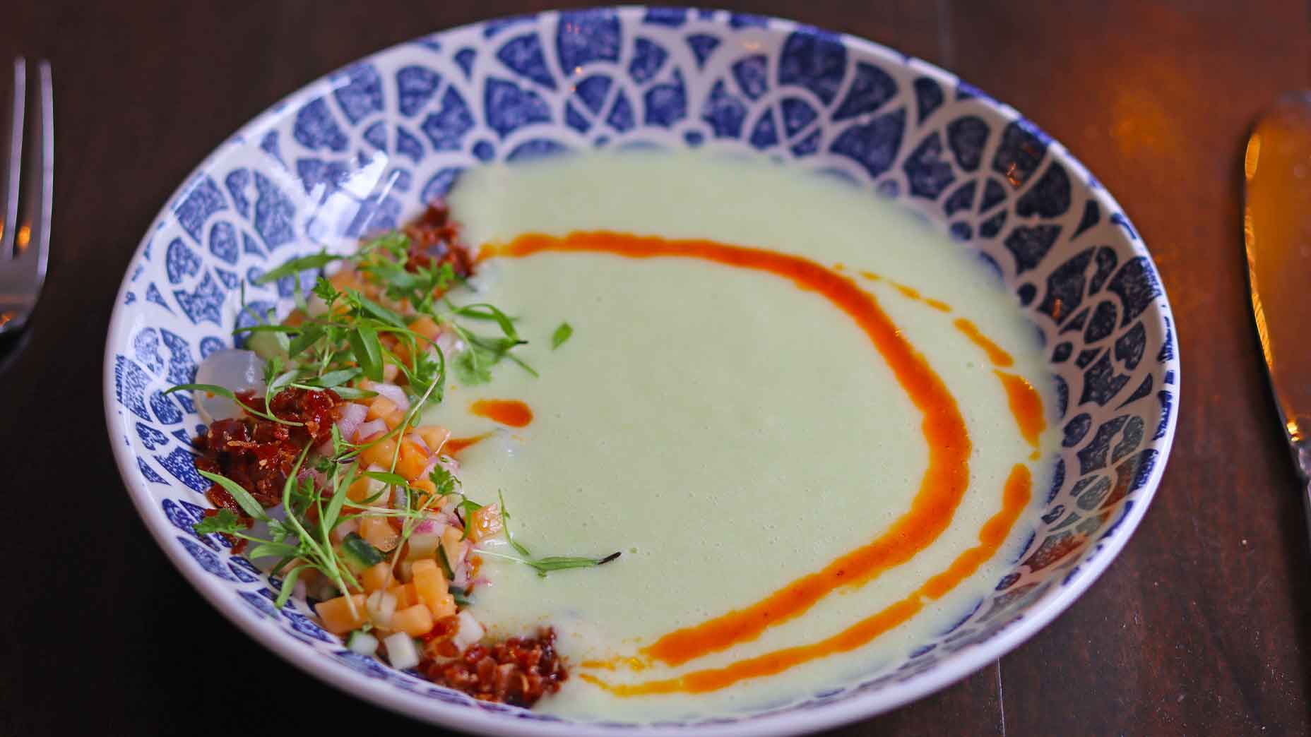 A patterned bowl with creamy green melon gazpacho, garnished with two orange sauce drizzles, diced vegetables, red grains, and microgreens on the side. A fork and knife are visible next to the bowl&mdash;perfect for enjoying your Virginia bites.