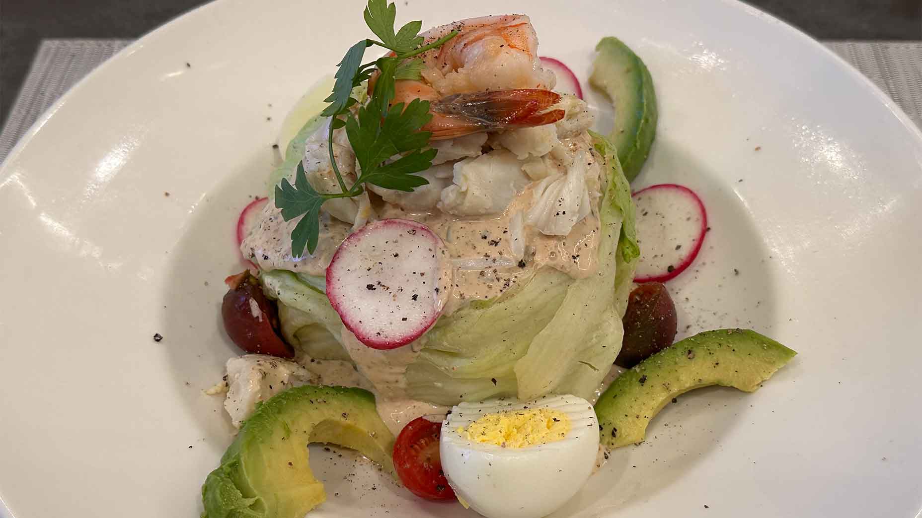A salad with iceberg lettuce, shrimp, crab, radish slices, cherry tomatoes, avocado, hard-boiled egg, and parsley, topped with dressing and black pepper, served on a white plate.