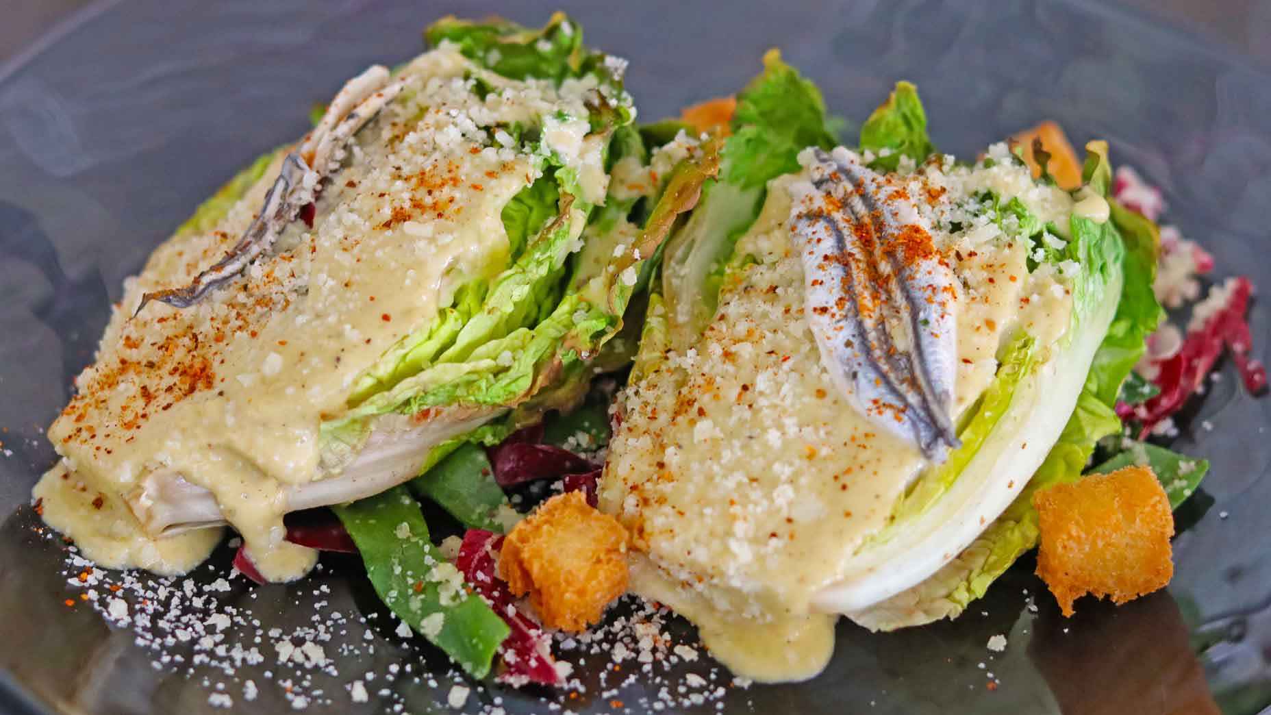 Two romaine lettuce wedges topped with Caesar dressing, anchovies, grated cheese, and paprika, served on a bed of mixed greens with croutons on a dark plate&mdash;a refreshing starter inspired by Virginia&rsquo;s fresh offerings and ideal to pair alongside local specialties such as bison ribeyes or a chilled melon gazpacho.