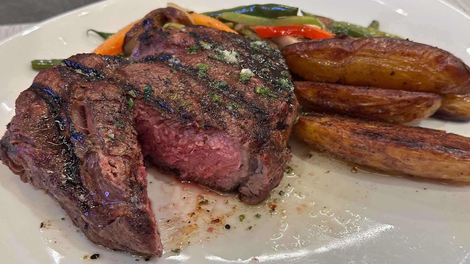A grilled steak cooked medium rare sits on a white plate, garnished with herbs. Roasted fingerling potatoes and saut&eacute;ed mixed vegetables are served on the side.