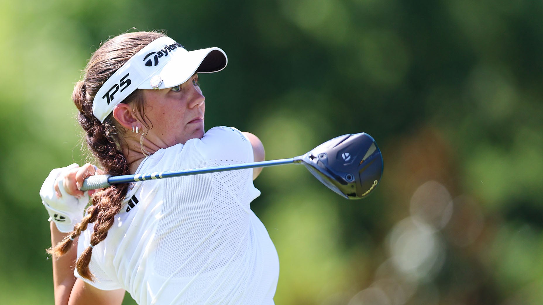 A female golfer wearing a white shirt and cap swings a driver golf club outdoors, with trees blurred in the background.