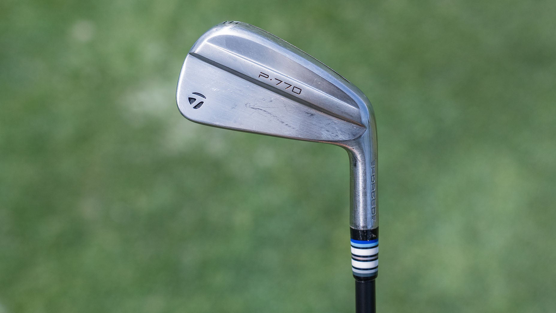 A close-up of a TaylorMade P-770 golf iron against a blurred green background, showing slight wear on the club head with a black, white, and blue striped ferrule.
