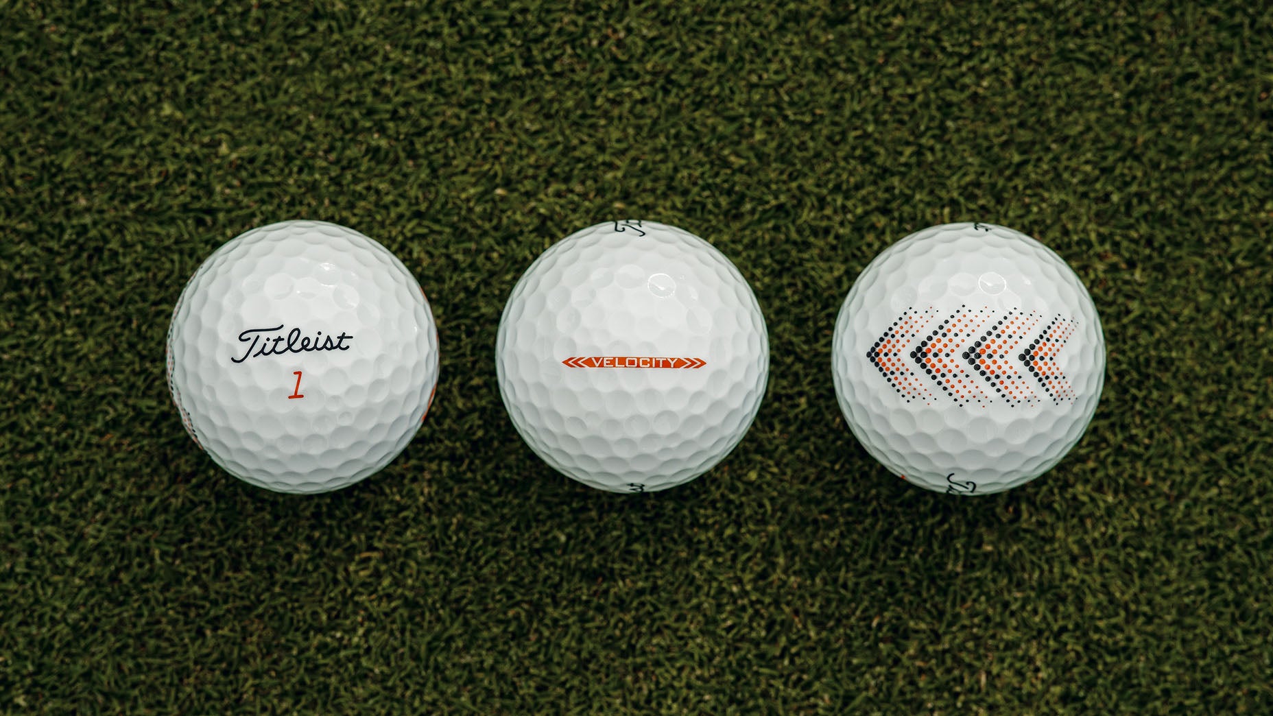Three white Titleist golf balls are lined up on green grass, each displaying different markings: the first shows the Titleist logo and a red number 1, the second says VELOCITY, and the third features red and black arrows.