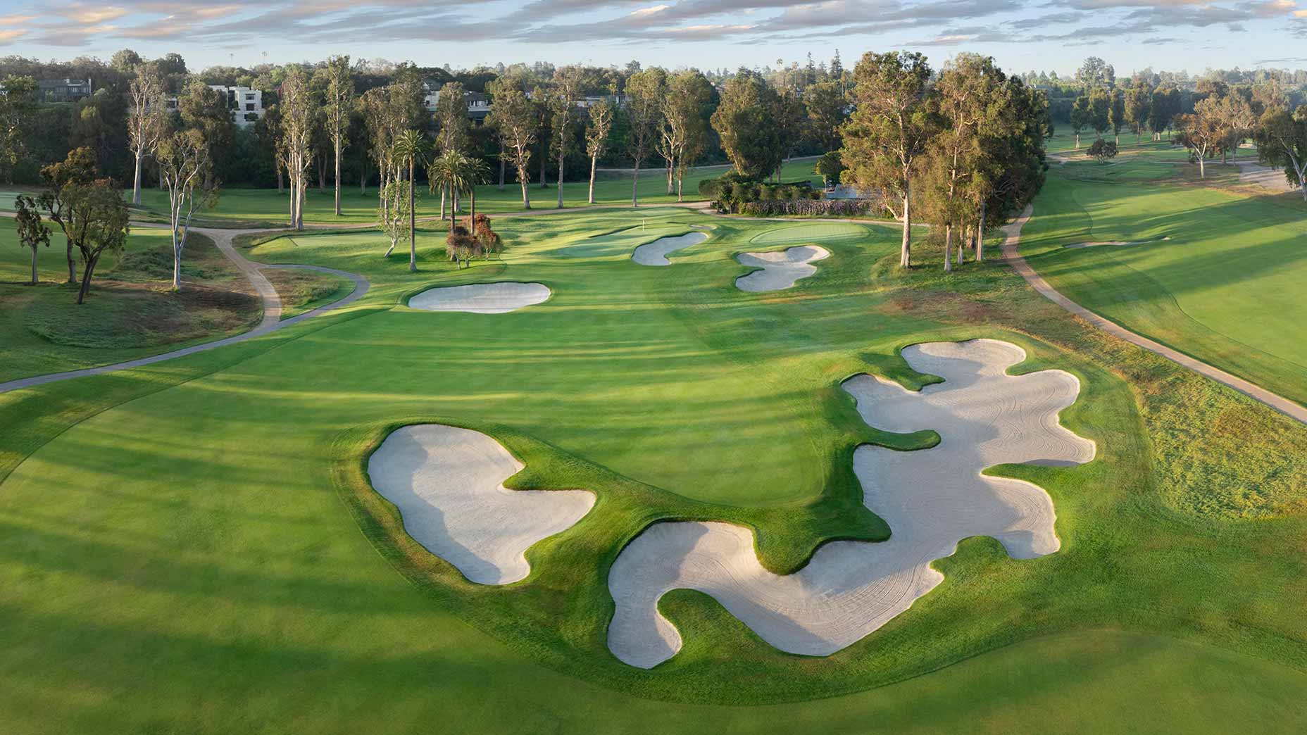Aerial view of a golf course reminiscent of Riviera, featuring lush green fairways, large sand bunkers, winding paths, and tall trees under a partly cloudy sky&mdash;perfect for hosting the U.S. Women's Open.