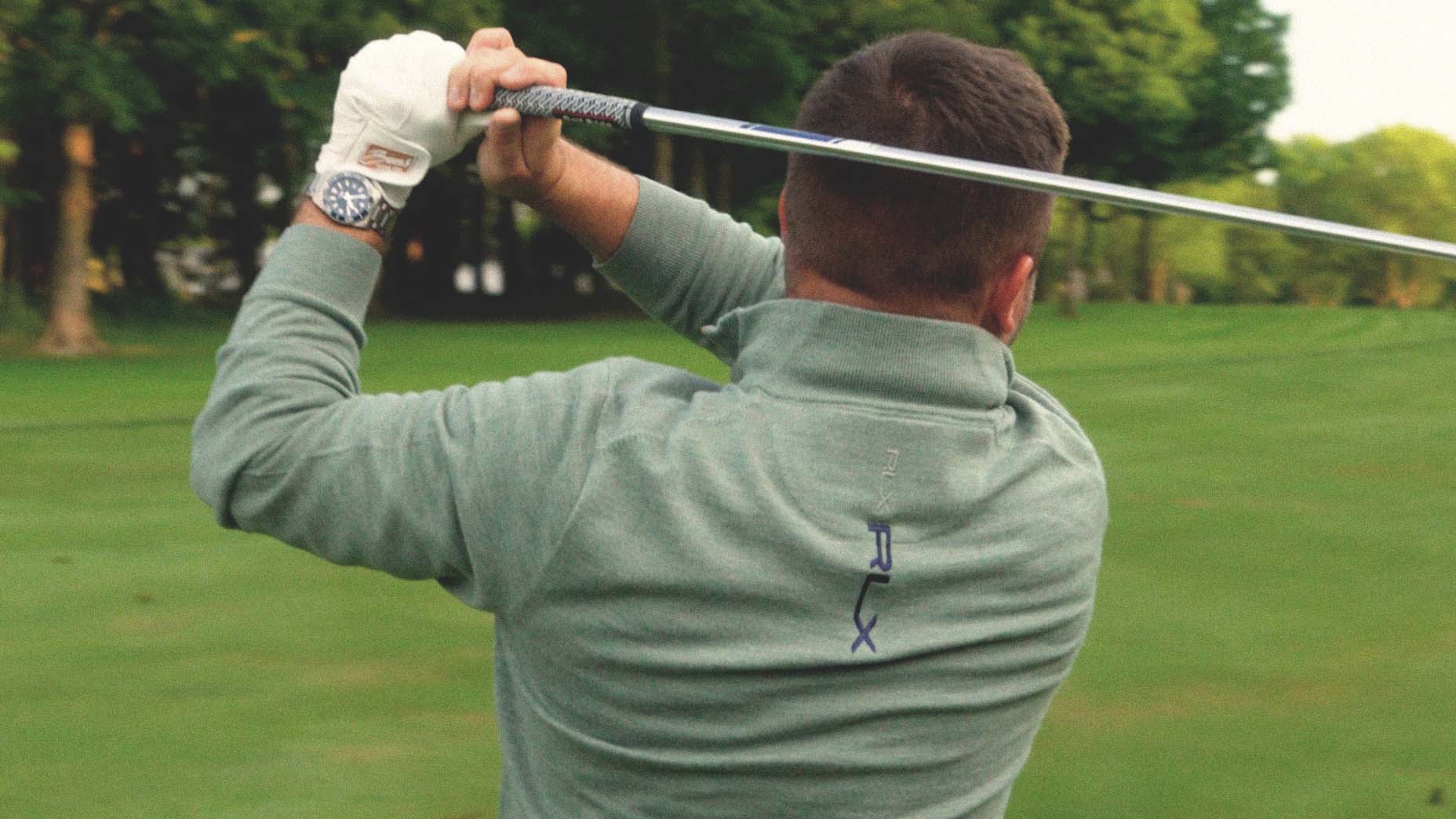 A person wearing a green jacket finishes a golf swing on a lush golf course, holding a club over their shoulder. Trees and green grass are visible in the background.