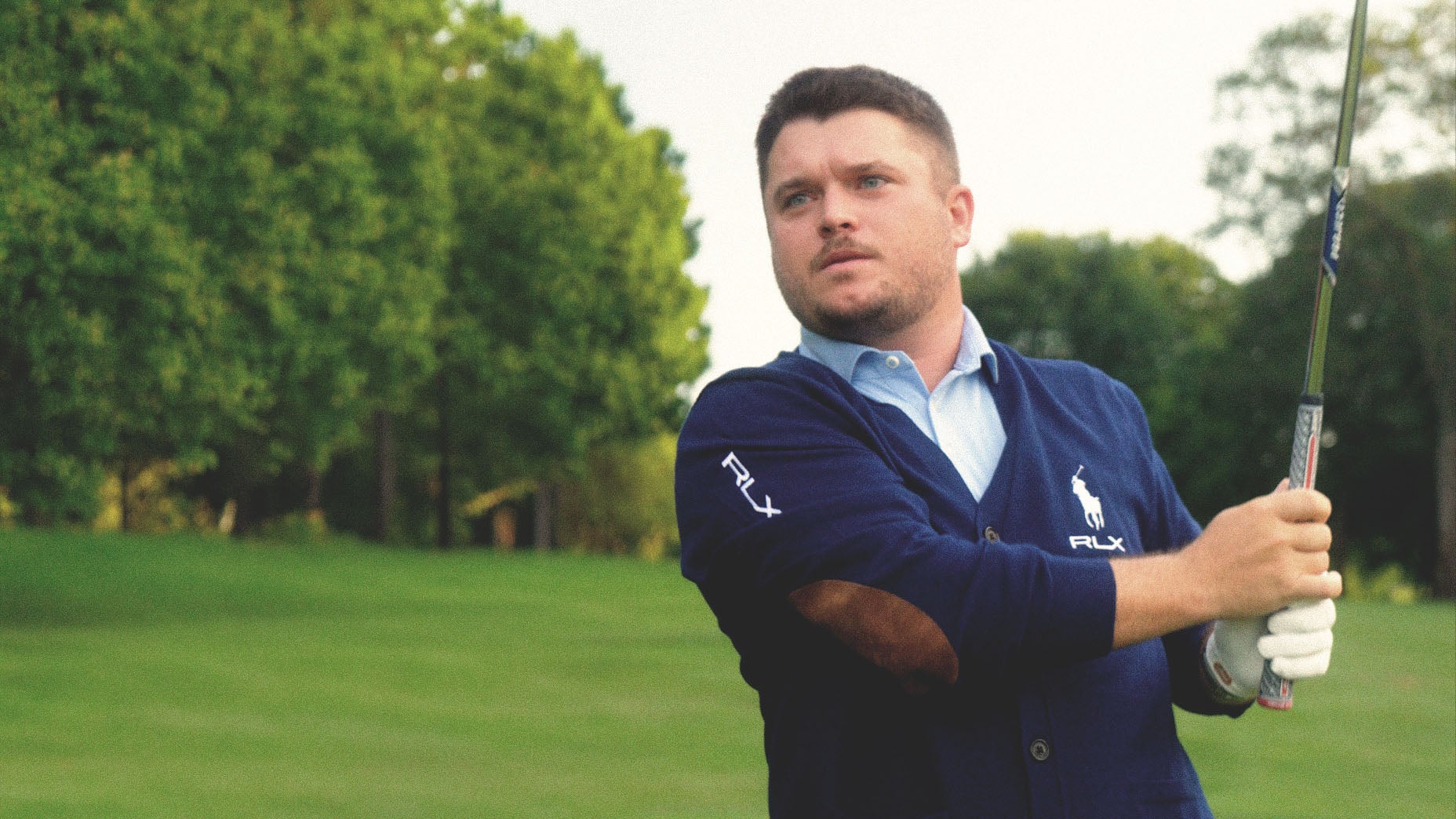 A man wearing a navy sweater and white golf glove holds a golf club, appearing to have just completed a swing on a green golf course with trees in the background.