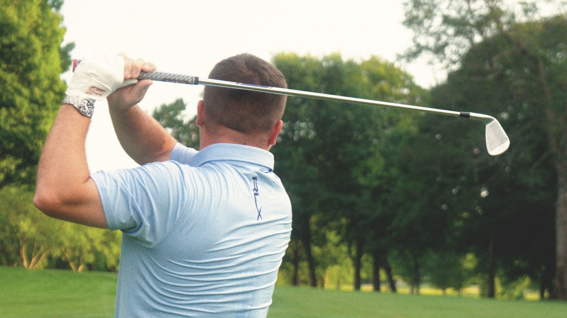 A man wearing a light blue shirt and golf glove follows through on a golf swing with a club, standing on a green fairway surrounded by trees.