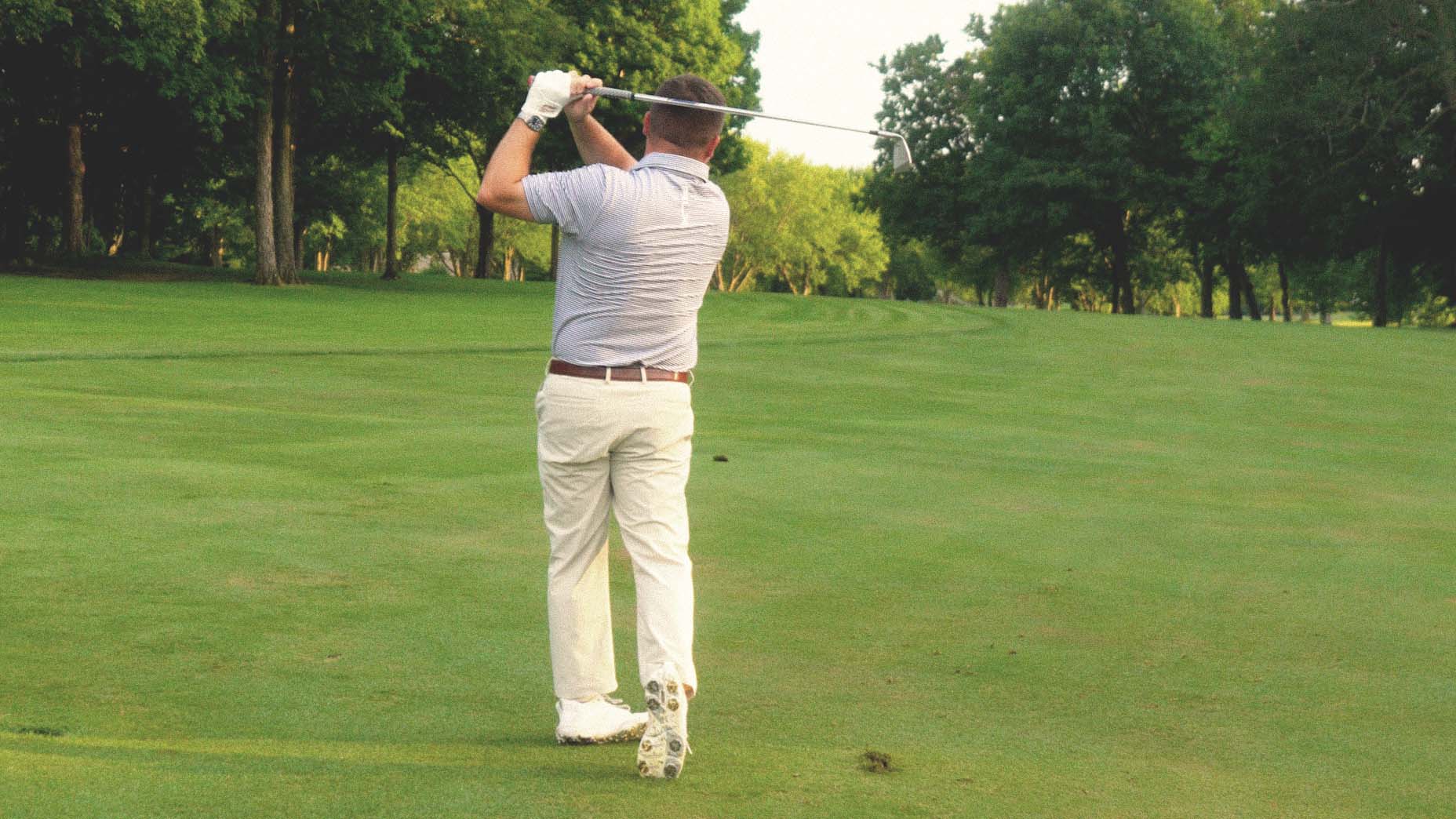 A man wearing a light shirt and pants swings a golf club on a green golf course, with trees and grass in the background.